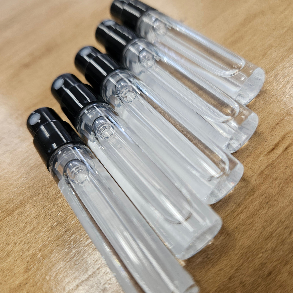 Five clear glass 2 ml perfume vials with black atomizers arranged diagonally on a light brown wooden surface. 