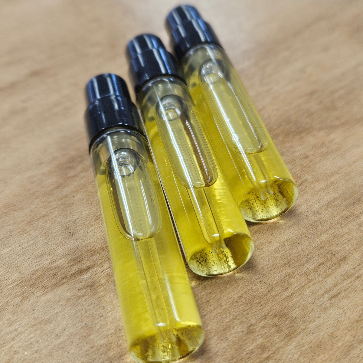 Three clear glass 2ml spray vials filled with yellow perfume and topped with black caps, arranged diagonally on a light brown wooden surface.