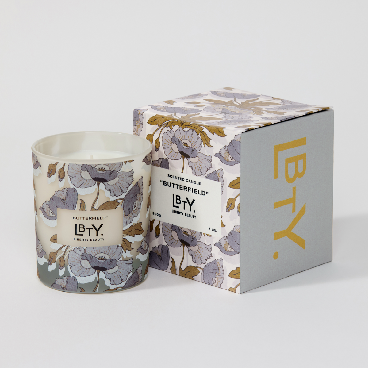 A scented candle in a floral-patterned glass jar sits beside its matching product box on a light gray background. It is the Butterfield candle by Liberty London. The notes are bergamot peel, cistus leaves, suede, violet, jasmine, patchouli steam, cedar leaf, amber, musk, vanilla.