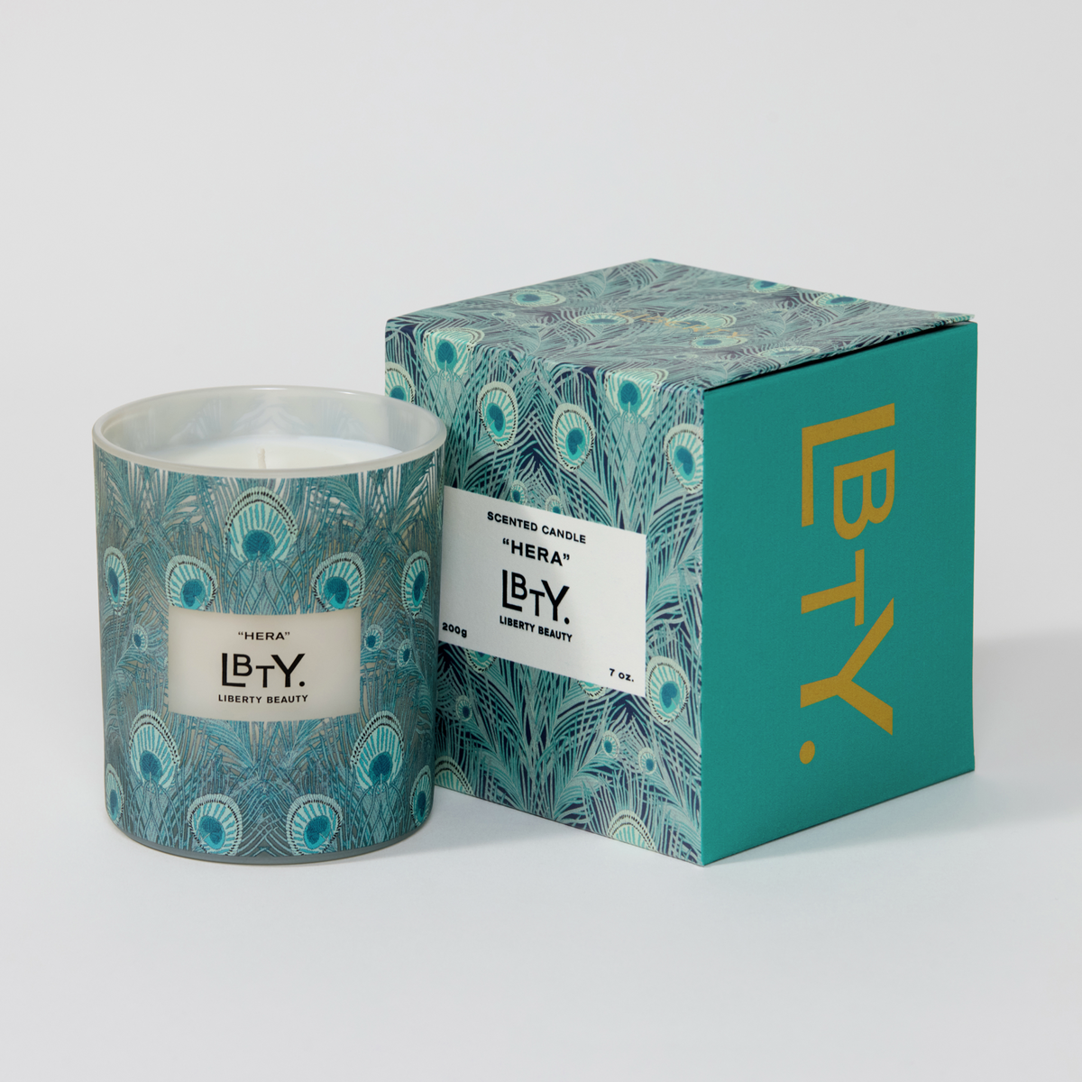 A scented candle in a glass jar with a teal and blue peacock feather pattern, shown next to its matching patterned box on a light gray background. The candle is Hera by Liberty London. Its notes include bartlett pear, freesia, citrus zest, golden amber, rose essence, benzoin, woodland moss, musk crystals, and dry woods. 